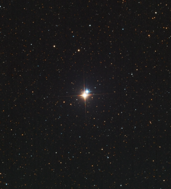 Two stars stand out against a starfield filled with numerous stars that appear as pinpricks of light.  The two stars are larger with 4-point diffraction spikes.  The star in front is a warm gold while the other star, one o'clock relative to the gold star, is a cool blue.